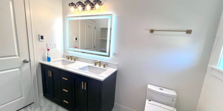 Upscale bathroom showcasing a dual-sink vanity with black shaker-style cabinet, gold hardware, marble countertops, and large illuminated mirror with LED backlighting. Includes white toilet with bidet seat and marble-patterned flooring — perfect for luxury design.