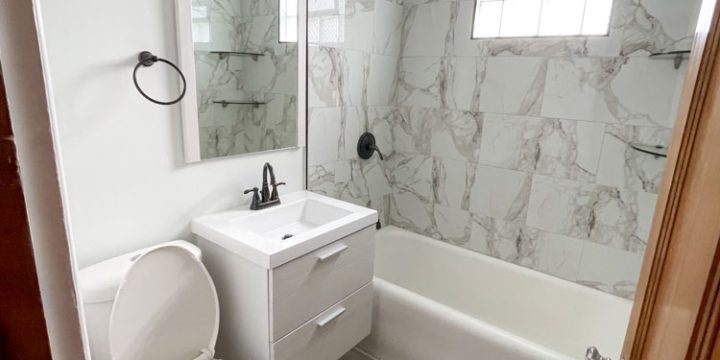 Transformed bathroom featuring white shaker-style vanity, marble-look tile walls and floor, white bathtub with chrome fixtures, and large mirror. Includes black hardware and recessed lighting — creating a clean, contemporary, and spa-like retreat.