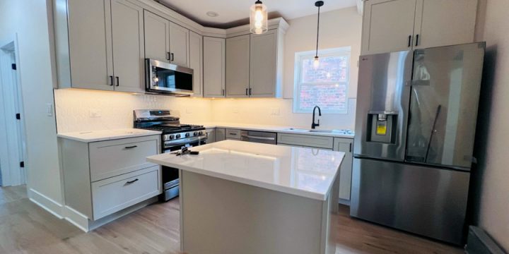 Bright, modern kitchen featuring light gray shaker-style cabinets, white marble countertops, stainless steel refrigerator and range, and large island with pendant lighting. Includes hardwood flooring and view to window — ideal for functional, elegant family living.