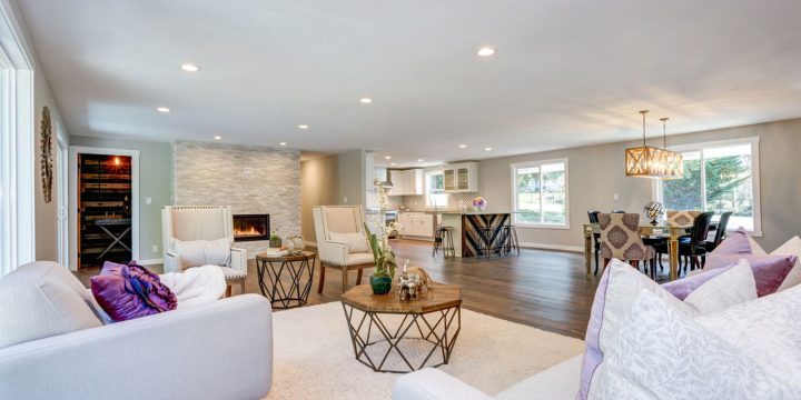 Spacious, updated living area featuring a stone fireplace, white sofas with purple accents, geometric coffee table, and view into kitchen with island and bar stools. Includes dining area with chandelier and large windows — creating a bright, cohesive, and inviting space.