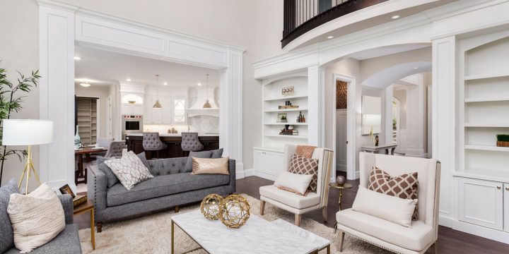 Elegant living space featuring gray sofa, cream armchairs, marble-top coffee table, and plush rug. Showcases white built-in shelves, arched doorways, and upper-level balcony with black railing. View into kitchen with dark cabinetry and pendant lighting — creating a grand, cohesive, and inviting interior.