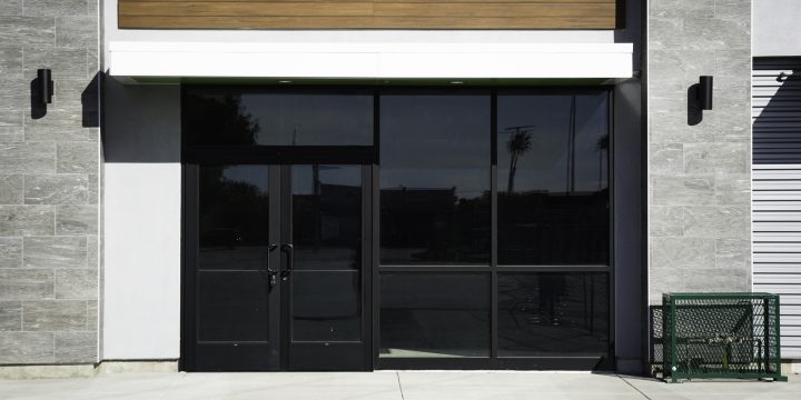 Contemporary storefront entrance featuring black-framed glass double doors, large windows, and modern facade with gray stone tile and horizontal wood siding. Includes black wall sconces and clean concrete sidewalk — projecting professionalism and upscale design.