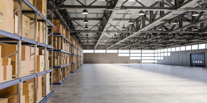 Spacious industrial warehouse featuring high ceilings with exposed steel trusses, pendant lighting, concrete flooring, and rows of blue metal shelving stocked with cardboard boxes. Large windows and roll-up doors provide natural light and access — ideal for commercial or logistics use.