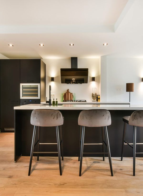 Sleek contemporary kitchen featuring a black island with white countertop, three gray velvet bar stools, recessed lighting, and wall-mounted sconces. Stainless steel appliances and light wood flooring complete the clean, inviting space ideal for calls to action.